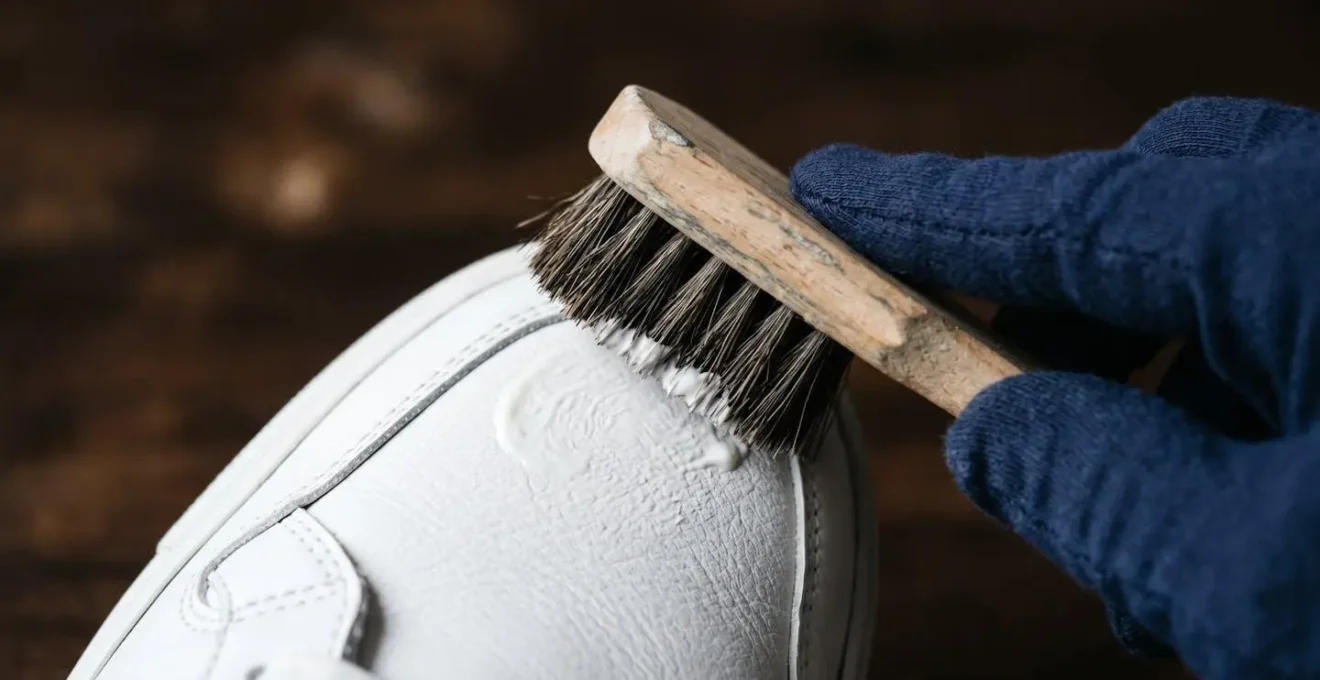 Gros plan sur des mains appliquant délicatement une crème d'entretien sur une sneaker en cuir blanc avec des outils professionnels